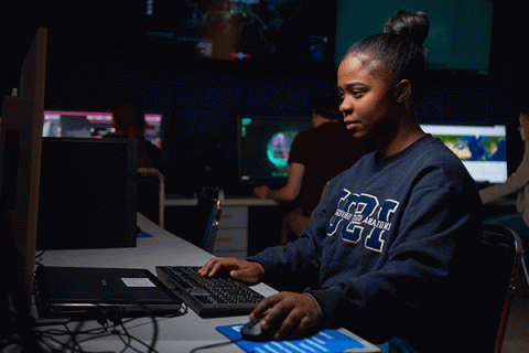 Female student of color with black hair in a bun and wearing a navy Indiana State University sweatshirt works on computer in a dark computer lab. Illuminated computer terminals and large-screen monitors are visible behind her.